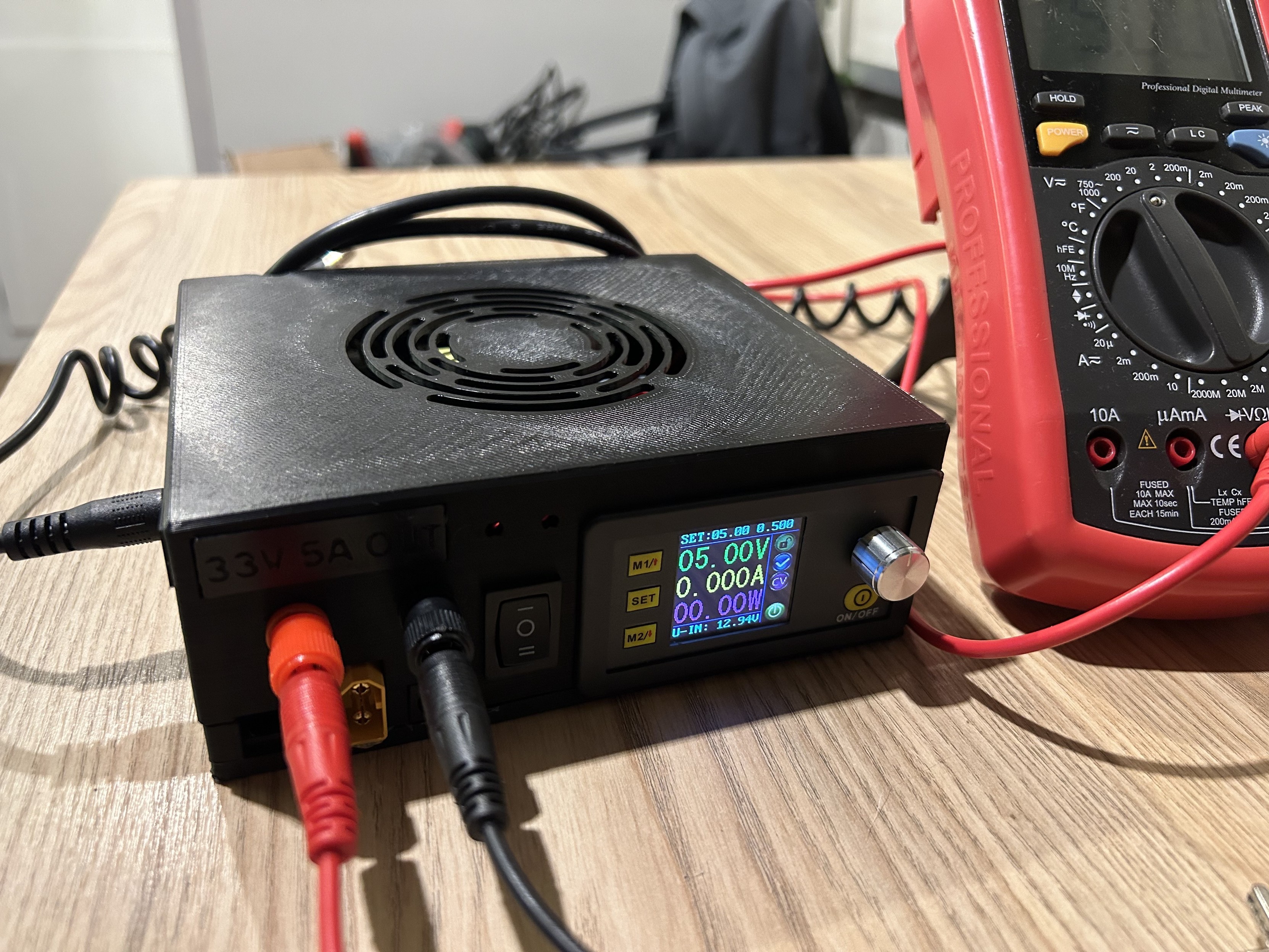 Zasilacz biurkowy i multimetr cyfrowy na drewnianym stole, z podłączonymi kablami.